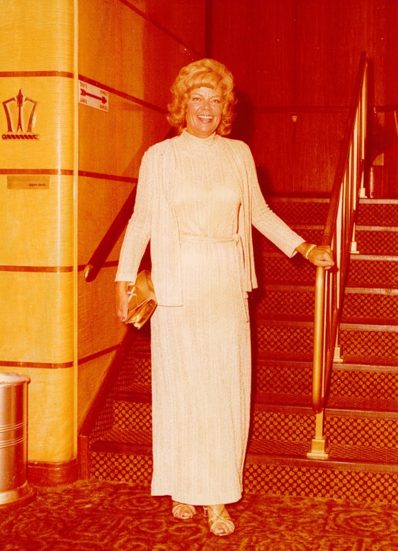A smiling woman wearing holiday dress stands with a clutch in her hand at the stairs.
