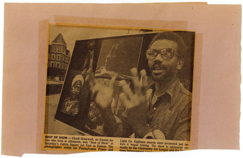 Newspaper clipping featuring a speaking man that stands in front of board with his artworks.
