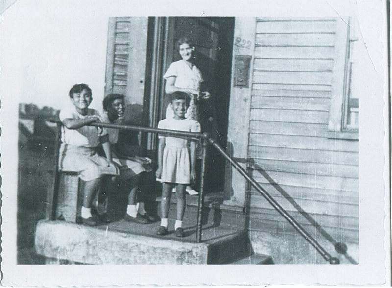 Three children at the top of a stoop. A woman is standing in the house's doorway.