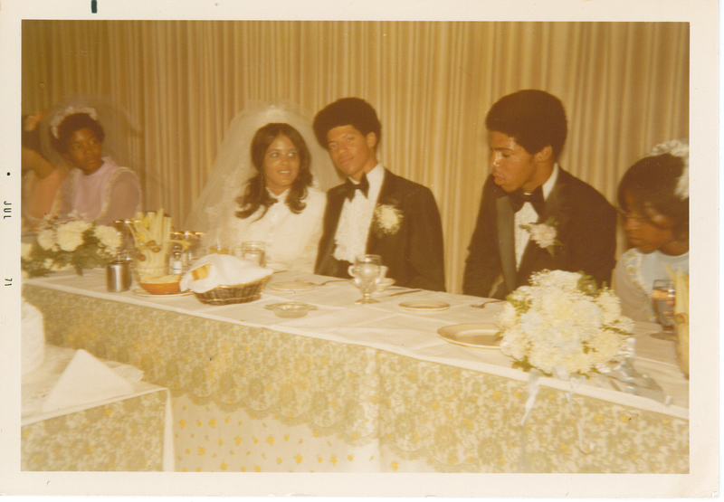 Multiple individuals dressed in formal wear sitting at a dining table. The presumed groom is making eye contact with the camera.