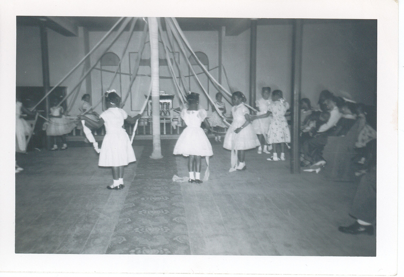 Young girls in white dresses dancing around a May Pole.