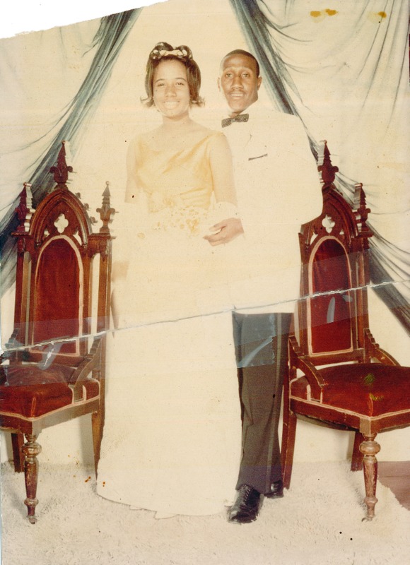 A man in a white jacket and a woman in a holiday dress standing in front of antique chairs.