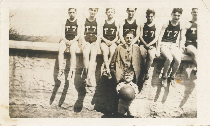 Seven boys sitting atop a wall. Each of them is wearing a jersey labeled "T7." A man in a suit stands in front of them.