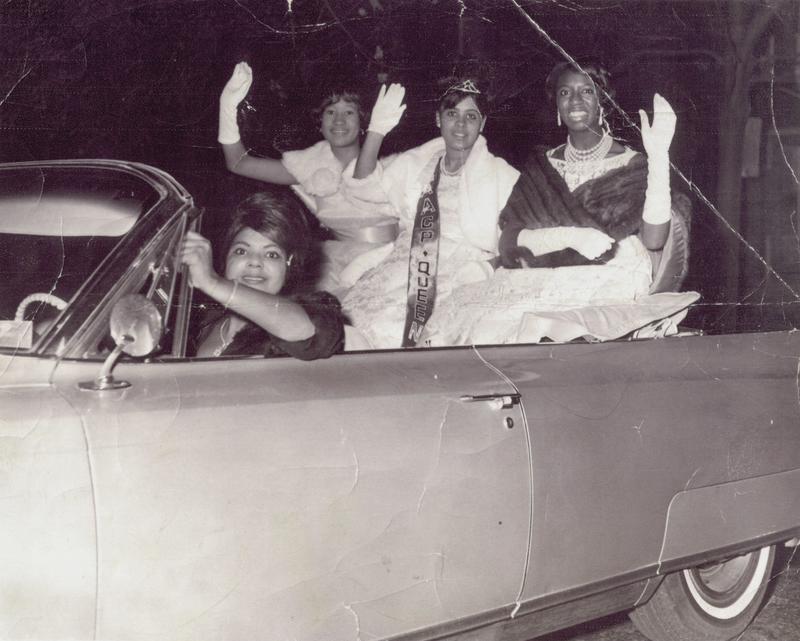 A group of women that wear formal dresses driving on a car and waving their hands.
