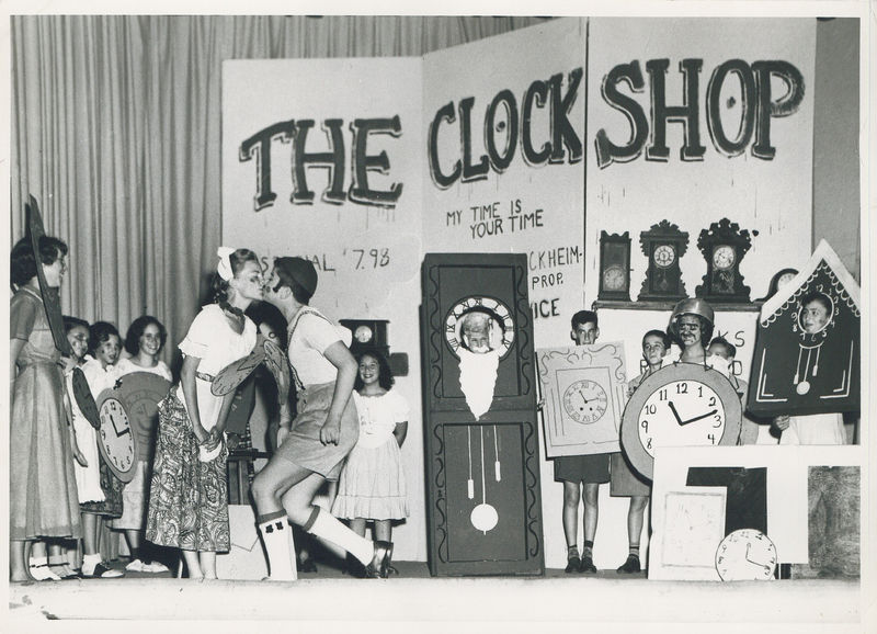 Multiple children observing two stage actors kissing. Each child is wearing a piece of paper with an illustration of a clock.