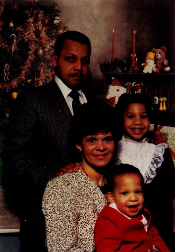 Husband and wife with two children wearing holiday dresses in front of a Christmas tree.