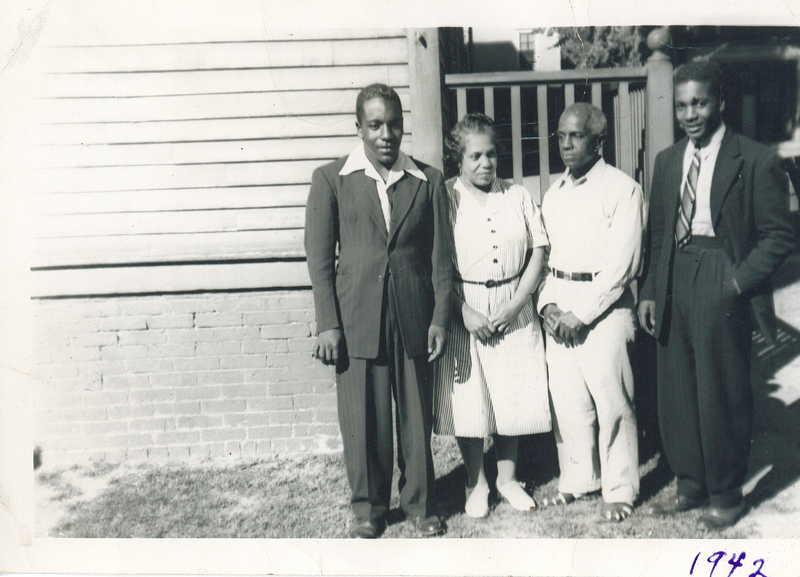 Two men in suits standing in front of the side of a house. They are flanking an older man and a woman.