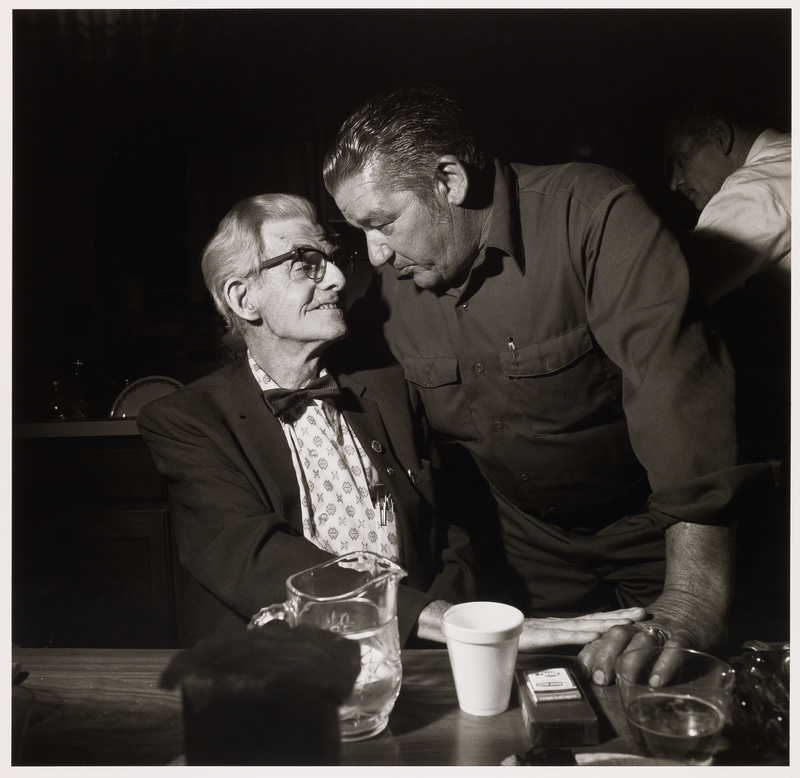 Two men looking at each other. The elderly man on the left, wearing glasses and a bowtie, is seated. The other man is younger and is standing with his right arm around the elderly man. Their hands touch on the table in front of them.