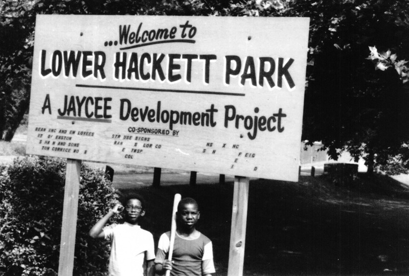 Two boys (one with baseball bat) standing under a sign.
