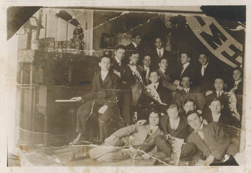 Several men sitting or standing. One is sitting at a piano.