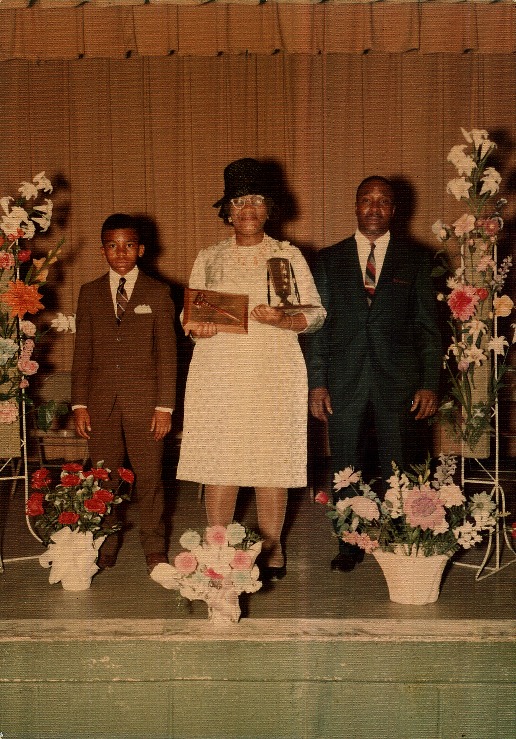A woman in a white dress and black hat stands with a gavel in her hands between a boy and a man in formal attires.