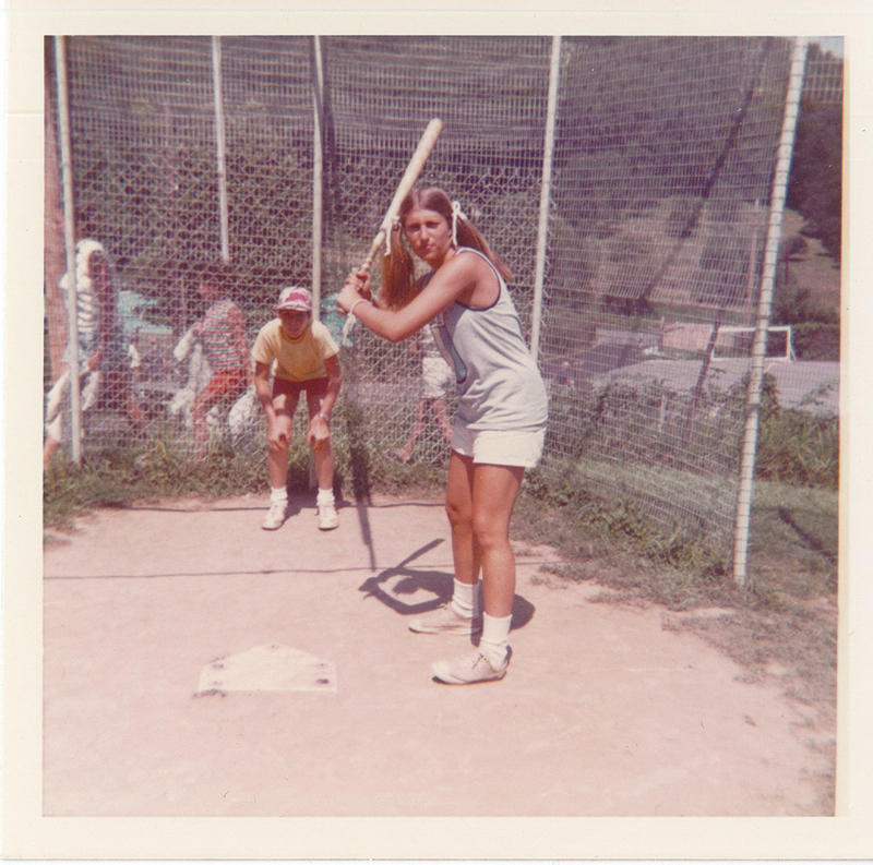 A young girl holding a bat to the right of home plate. A boy with his hands on his knees stands behind the plate.