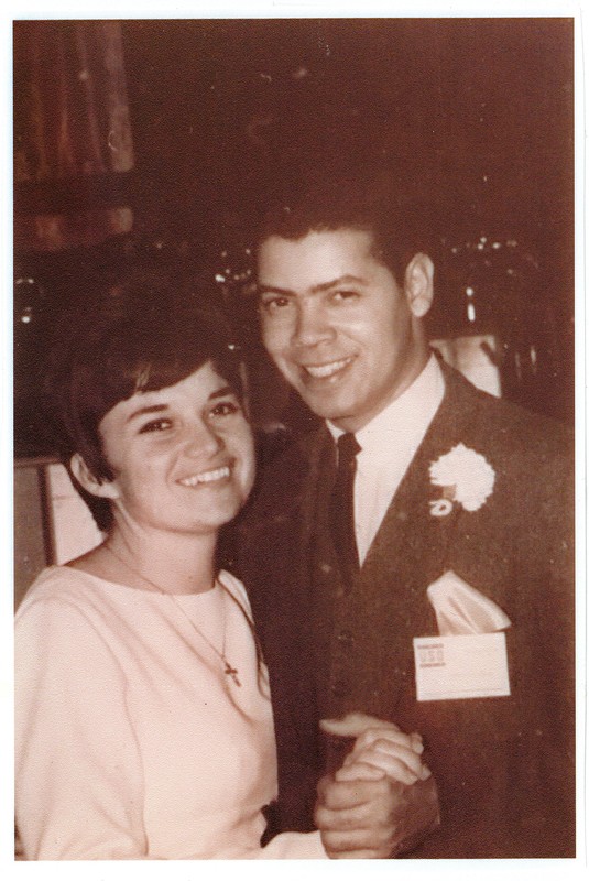 A man in a suit holding hands with a woman in a white dress. Both are smiling.