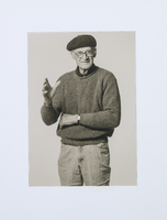 An elderly man with glasses and a beret. He has one arm crossed across his stomach and is gesticulating with the other hand. 