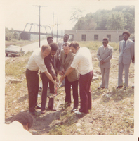 Four men holding a shovel. Other men are standing behind them and observing.