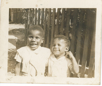 Two children standing in front of a wooden fence gate. Their arms are wrapped around each other.