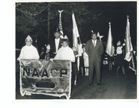 A group of individuals marching. Some are holding various flags. Two women at the front of the group hold a sign indicating the Easton branch of the NAACP.