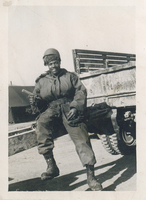 A man in military fatigues sitting on top of a drawbar. He is holding a handgun.