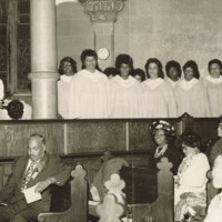 A group of female choir singers in white dresses and church visitors sitting on the benches.
