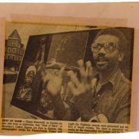 Newspaper clipping featuring a speaking man that stands in front of board with his artworks.