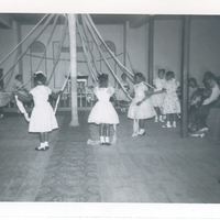 Young girls in white dresses dancing around a May Pole.