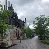 A park with brick paving. The steel mill can be seen to the left. Metal sculptures are scattered about the park.
