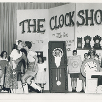 Multiple children observing two stage actors kissing. Each child is wearing a piece of paper with an illustration of a clock.