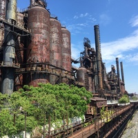 Rusted boilers against a partly cloudy sky. Trees have begun to overtake various areas of the plant.
