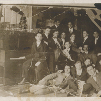 Several men sitting or standing. One is sitting at a piano.