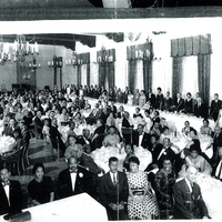 A large group of individuals gathered in a hall. All are dressed in evening wear. Candle chandeliers hang from the ceiling.